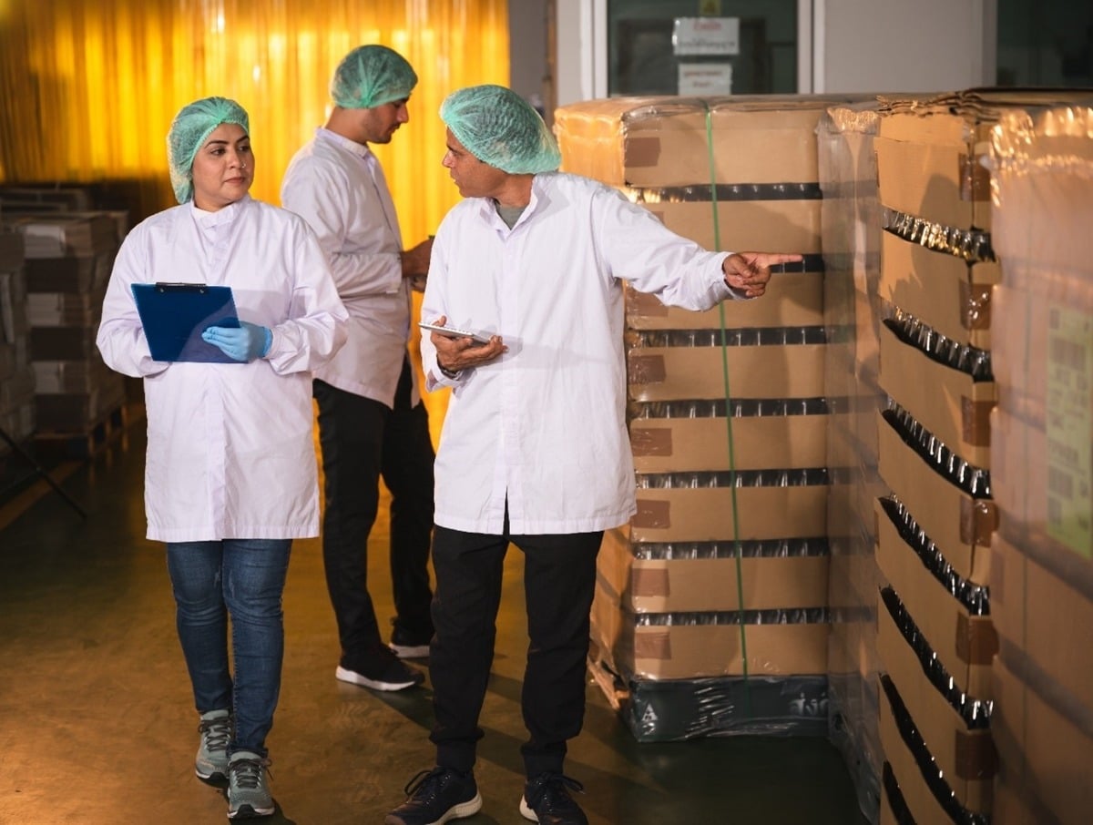 Trois employés en blouses blanches inspectent des cartons emballés dans un entrepôt industriel.